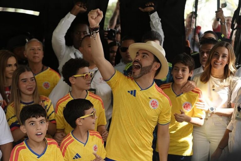 Man in a white shirt waving from a stage to a cheering crowd holding yellow, blue and red flags at night.