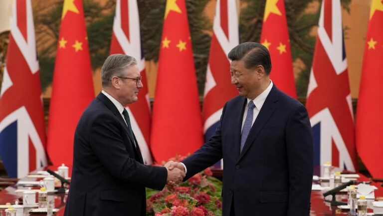 Two men in dark suits shaking hands in front of British and Chinese flags in a formal hall.