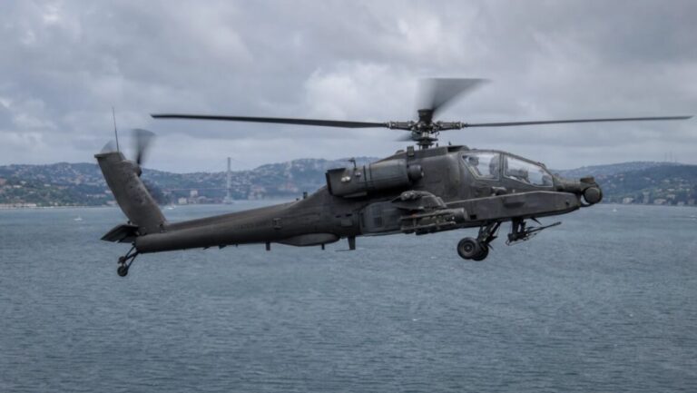 Dark gray attack helicopter with a slender fuselage and stub wings flying low over desert terrain.